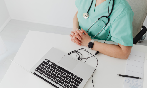 Lekarz w zielonym stroju medycznym ze stetoskopem siedzący przy biurku przed laptopem, symbolizujący konsultację medyczną online w zakresie diagnostyki i leczenia choroby Leśniowskiego-Crohna.