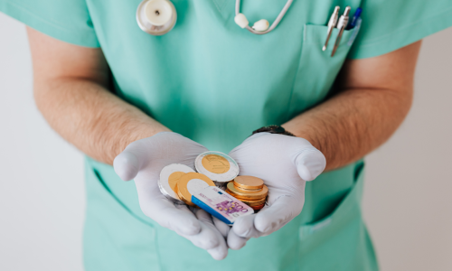 Pracownik medyczny w zielonym stroju i jednorazowych rękawiczkach trzymający w dłoniach monety i banknot, ilustrujący temat wynagrodzenia i finansowania udziału w badaniach klinicznych.