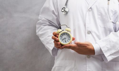 Lekarz w białym fartuchu ze stetoskopem trzymający w dłoniach mały zegar, symbolizujący czas trwania badań klinicznych oraz oczekiwanie na efekty terapii.